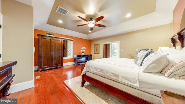 bedroom featuring baseboards, visible vents, wood finished floors, a tray ceiling, and multiple windows
