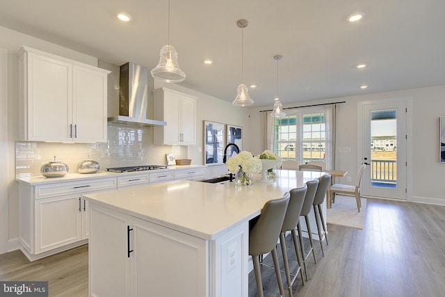 kitchen with stainless steel gas stovetop, white cabinetry, sink, wall chimney range hood, and a center island with sink