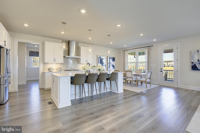 kitchen featuring an island with sink, pendant lighting, wall chimney range hood, and white cabinets