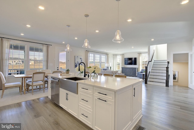 kitchen featuring white cabinets, hanging light fixtures, stainless steel dishwasher, a center island with sink, and light hardwood / wood-style flooring