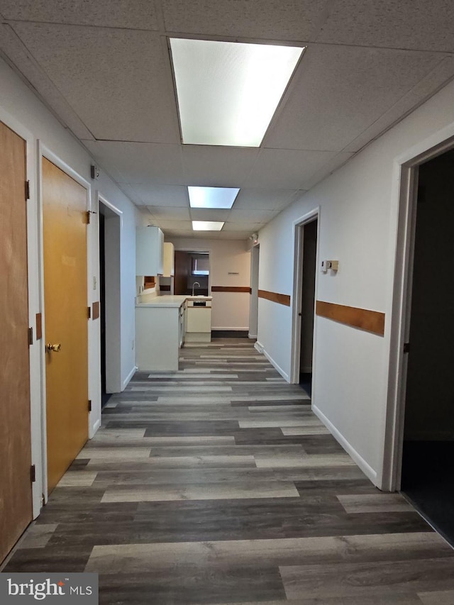 hallway with dark wood-style flooring, a drop ceiling, a sink, and baseboards