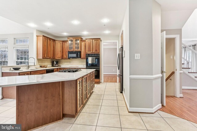 kitchen featuring light tile patterned floors, brown cabinetry, a kitchen island, glass insert cabinets, and black appliances