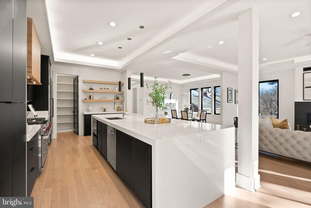 kitchen with sink, hanging light fixtures, a center island with sink, a tray ceiling, and light hardwood / wood-style floors