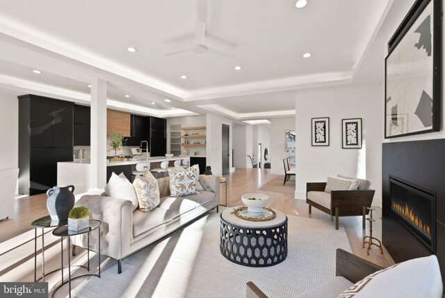 living room featuring a raised ceiling, ceiling fan, a large fireplace, and light wood-type flooring