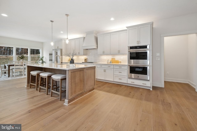 kitchen featuring pendant lighting, a kitchen island with sink, double oven, white cabinets, and custom exhaust hood