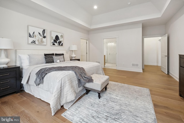 bedroom with ensuite bath, a raised ceiling, and light wood-type flooring