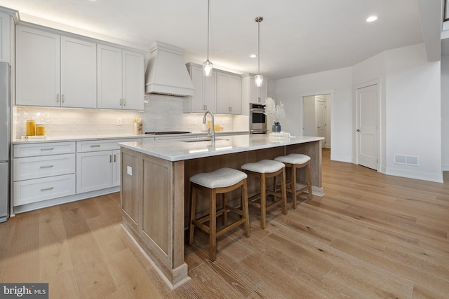 kitchen with premium range hood, pendant lighting, sink, a center island with sink, and light hardwood / wood-style flooring