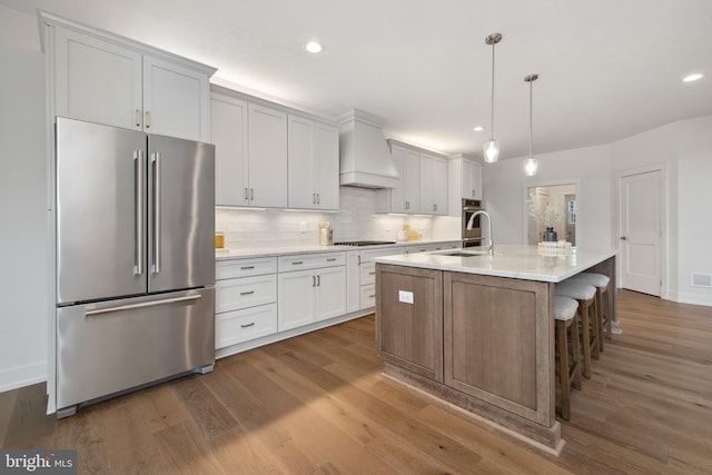 kitchen featuring premium range hood, an island with sink, white cabinets, hanging light fixtures, and high end fridge