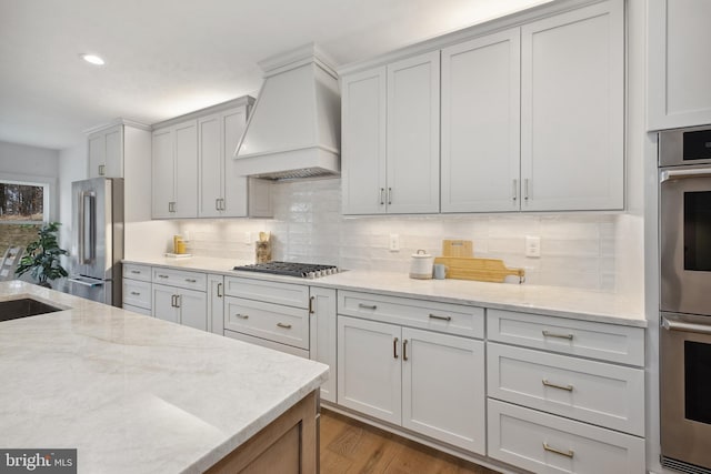 kitchen with sink, backsplash, stainless steel appliances, light stone countertops, and custom range hood