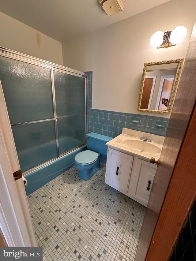 bathroom featuring bath / shower combo with glass door, toilet, a wainscoted wall, vanity, and tile walls