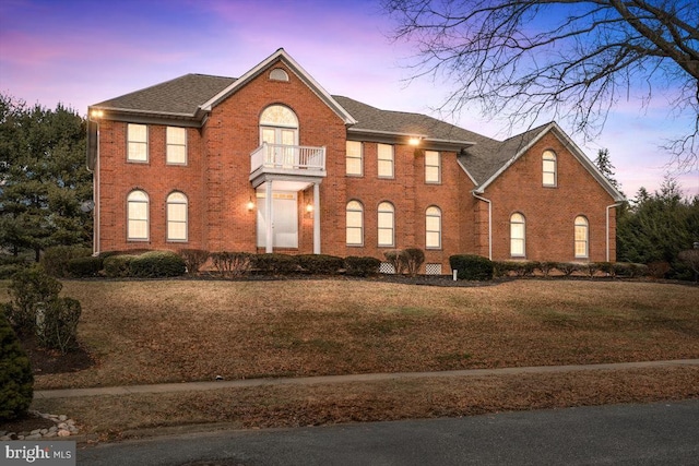 colonial house with brick siding, a balcony, a front lawn, and roof with shingles