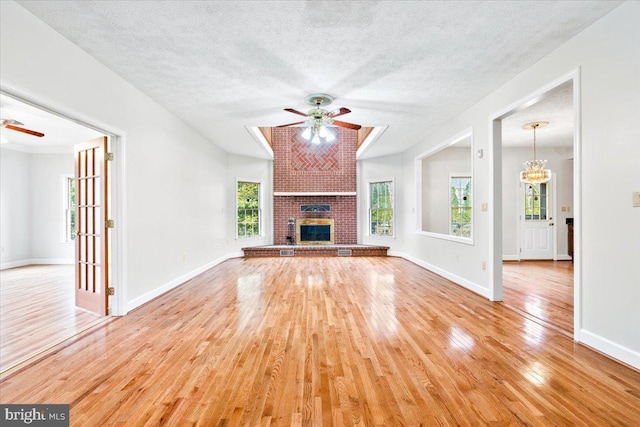 unfurnished living room with ceiling fan with notable chandelier, a brick fireplace, light wood-style flooring, and baseboards