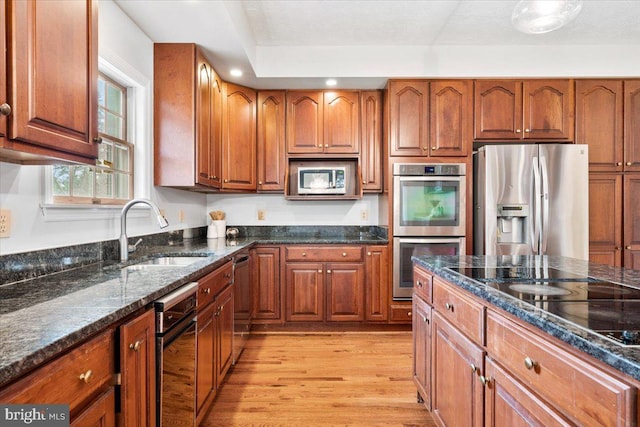 kitchen with light wood-style flooring, appliances with stainless steel finishes, brown cabinets, dark stone countertops, and a sink