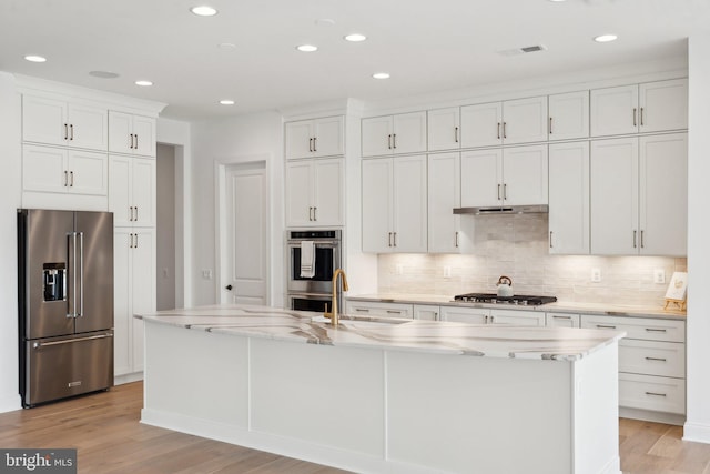 kitchen featuring appliances with stainless steel finishes, white cabinets, decorative backsplash, light stone counters, and a center island with sink