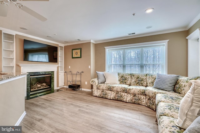 living room featuring built in features, ceiling fan, a high end fireplace, light hardwood / wood-style floors, and crown molding