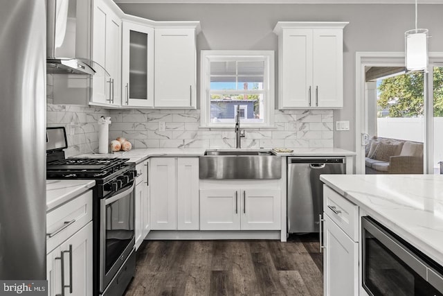 kitchen featuring wall chimney exhaust hood, sink, hanging light fixtures, appliances with stainless steel finishes, and white cabinets