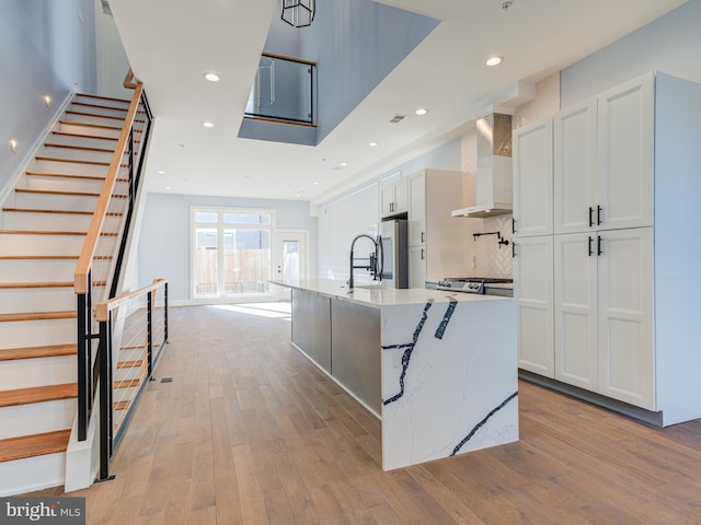 kitchen featuring white cabinetry, wall chimney exhaust hood, stainless steel refrigerator, light stone countertops, and a kitchen island with sink