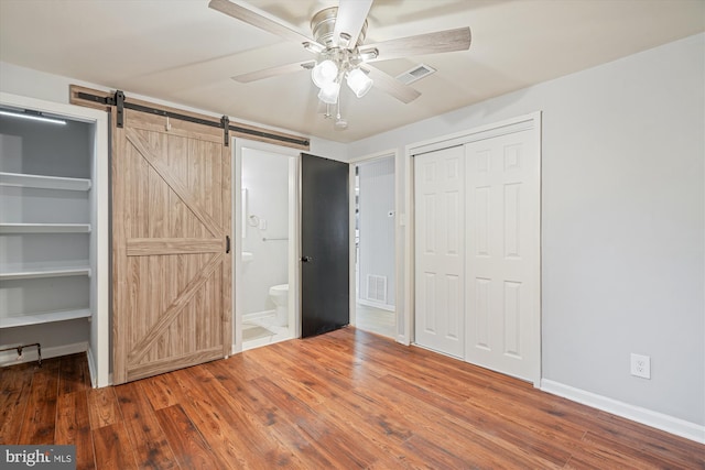 unfurnished bedroom featuring hardwood / wood-style flooring, ceiling fan, a barn door, ensuite bath, and a closet