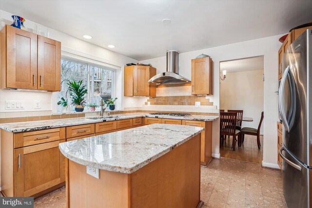 kitchen with wall chimney exhaust hood, a kitchen island, light stone counters, stainless steel appliances, and a sink