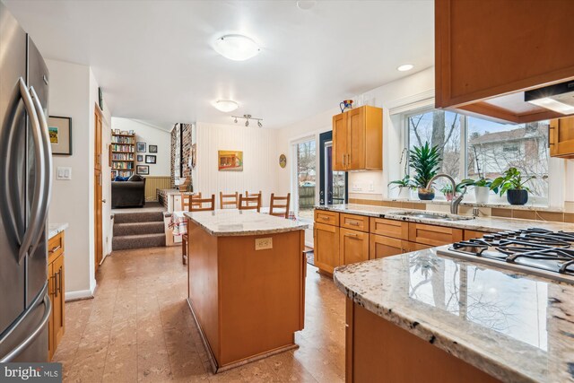 kitchen with brown cabinetry, a center island, light stone countertops, stainless steel appliances, and a sink