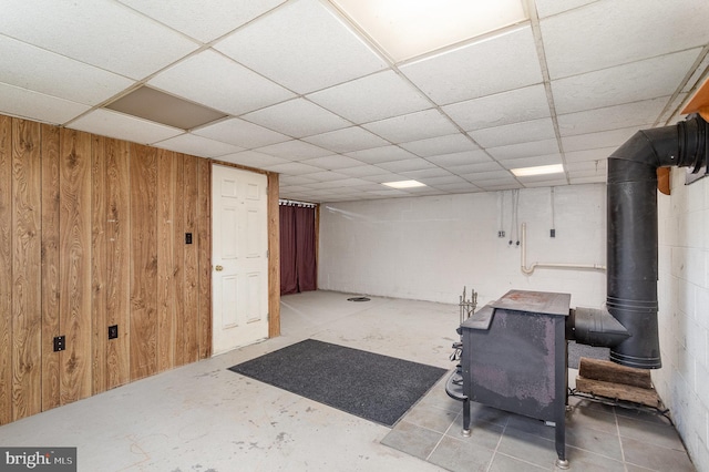 basement with wooden walls, a drop ceiling, and a wood stove