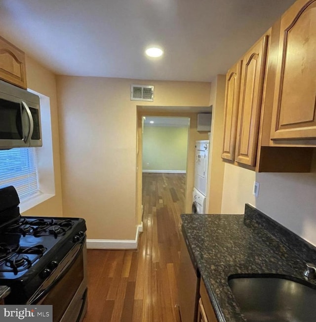 kitchen featuring appliances with stainless steel finishes, sink, dark stone countertops, dark hardwood / wood-style flooring, and stacked washer and clothes dryer