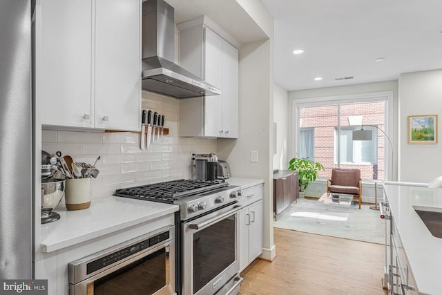 kitchen featuring stainless steel gas stove, white cabinets, wall chimney exhaust hood, light stone counters, and light hardwood / wood-style flooring