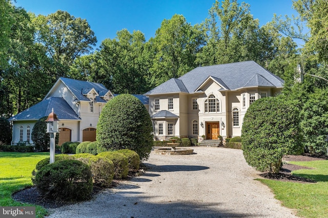 french country home featuring a garage, a front lawn, gravel driveway, and stucco siding