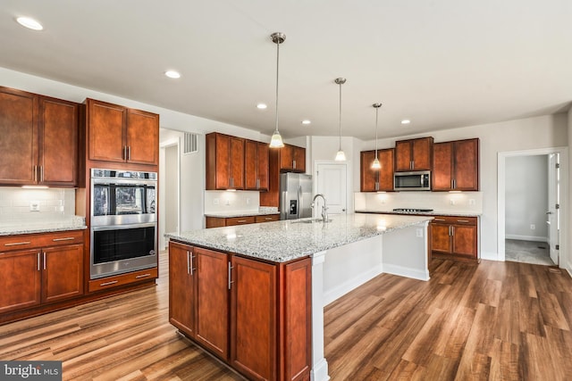 kitchen featuring hardwood / wood-style flooring, appliances with stainless steel finishes, a kitchen island with sink, hanging light fixtures, and light stone countertops