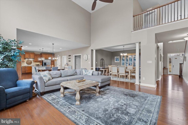 living area featuring hardwood / wood-style flooring, baseboards, a towering ceiling, ornate columns, and ceiling fan with notable chandelier