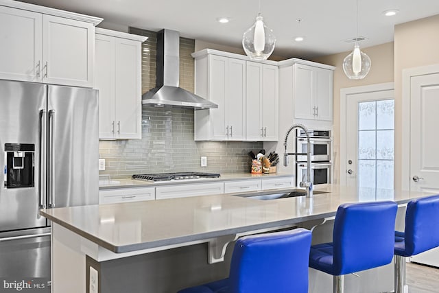 kitchen with white cabinetry, hanging light fixtures, stainless steel appliances, a center island with sink, and wall chimney exhaust hood