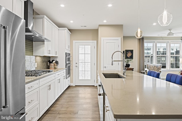 kitchen featuring wall chimney exhaust hood, sink, hanging light fixtures, an island with sink, and stainless steel appliances