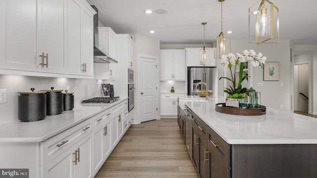 kitchen with appliances with stainless steel finishes, white cabinetry, hanging light fixtures, a kitchen island with sink, and wall chimney exhaust hood