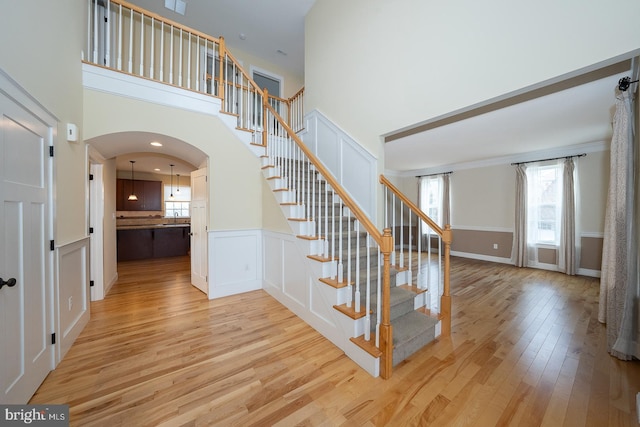 stairs featuring arched walkways, a decorative wall, wood finished floors, a towering ceiling, and wainscoting