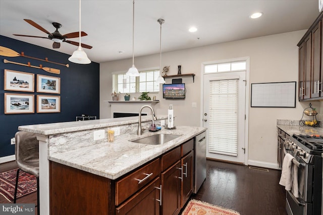 kitchen featuring sink, appliances with stainless steel finishes, dark brown cabinets, light stone countertops, and an island with sink