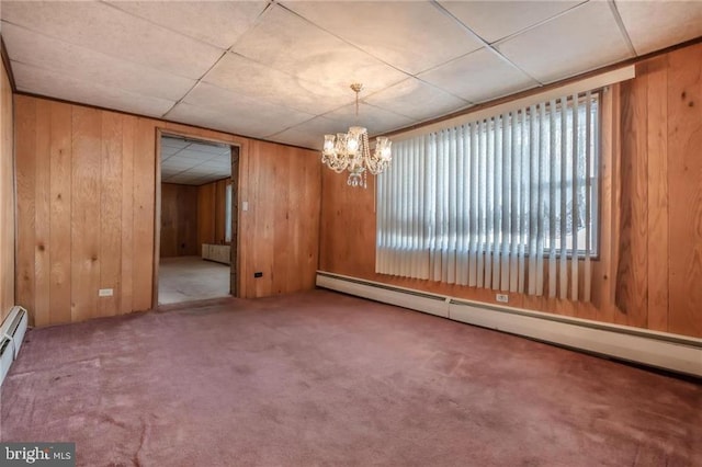 carpeted empty room featuring an inviting chandelier, a baseboard radiator, a drop ceiling, and wood walls