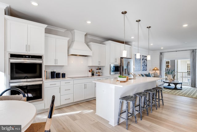 kitchen with a center island with sink, stainless steel appliances, light countertops, custom range hood, and white cabinets