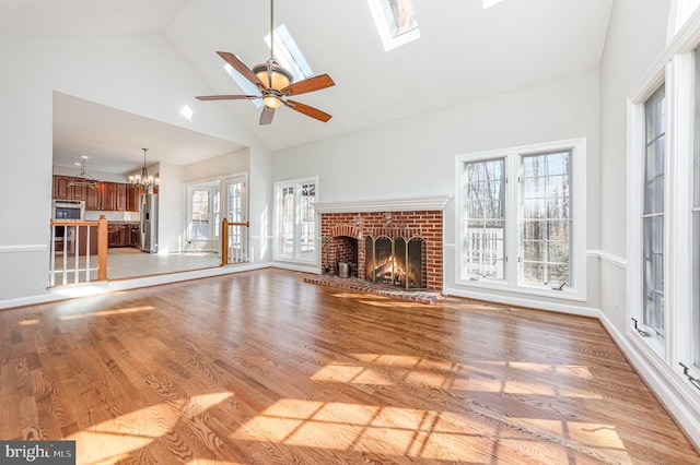 unfurnished living room with a skylight, light wood-style floors, a fireplace, high vaulted ceiling, and ceiling fan with notable chandelier