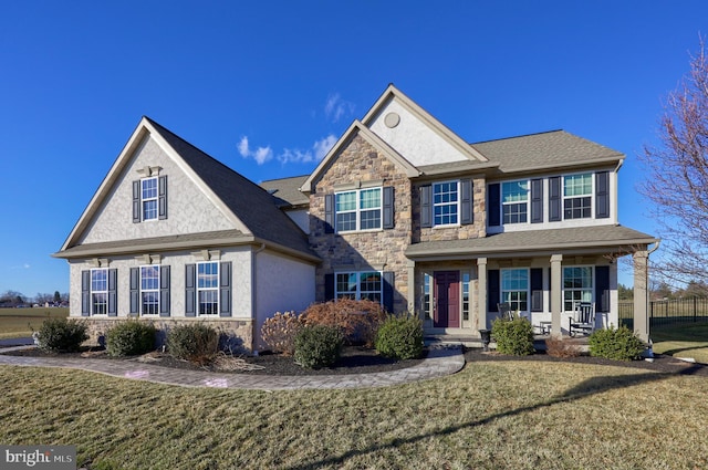 view of front facade with roof with shingles, a front lawn, stone siding, covered porch, and stucco siding