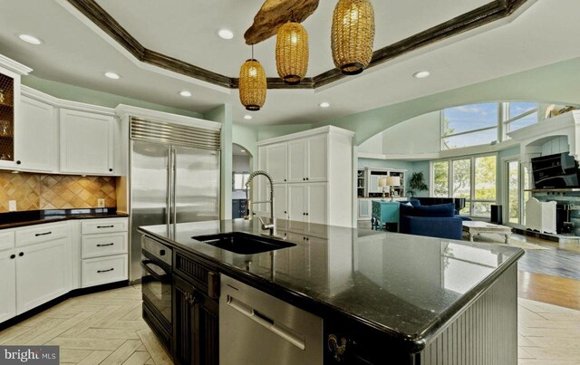 kitchen featuring sink, dark stone countertops, appliances with stainless steel finishes, a raised ceiling, and a kitchen island with sink