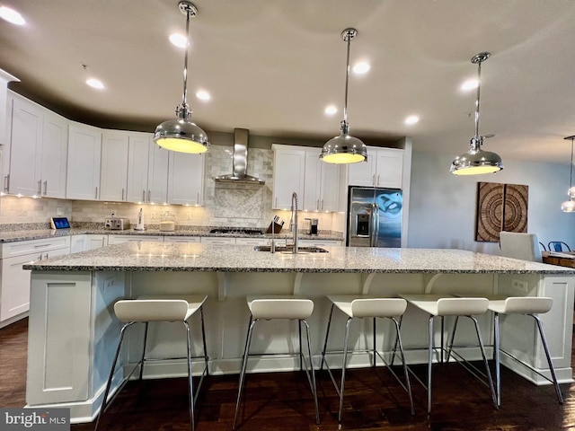 kitchen with a sink, a large island, stainless steel refrigerator with ice dispenser, backsplash, and wall chimney exhaust hood