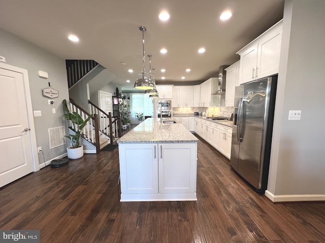 kitchen with wall chimney range hood, stainless steel appliances, light stone countertops, white cabinets, and a center island with sink