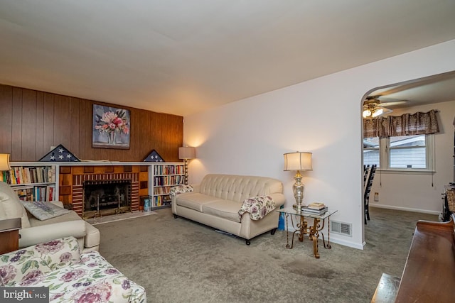 carpeted living room with a brick fireplace, wooden walls, and ceiling fan