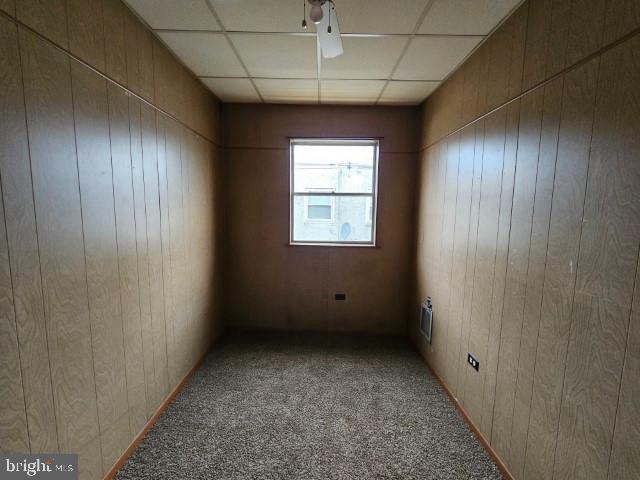 empty room featuring a drop ceiling, wooden walls, and carpet