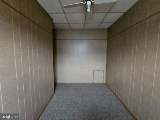 empty room featuring a drop ceiling, carpet floors, ceiling fan, and wood walls