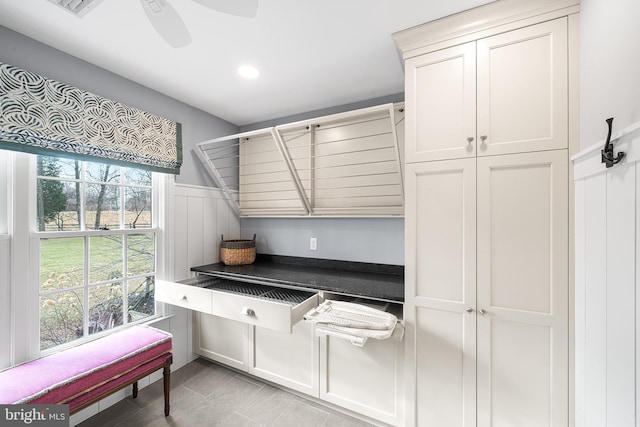 kitchen with a wainscoted wall, dark countertops, a decorative wall, a ceiling fan, and white cabinetry