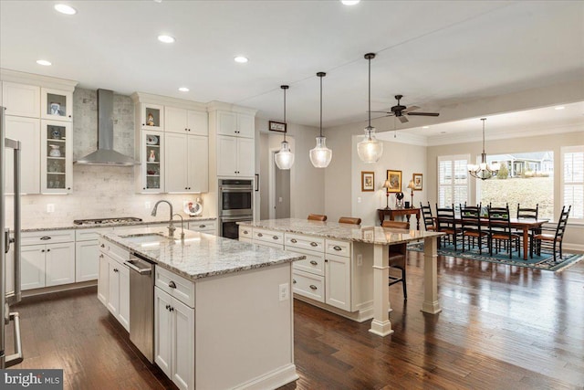 kitchen featuring a center island with sink, glass insert cabinets, appliances with stainless steel finishes, wall chimney range hood, and a sink