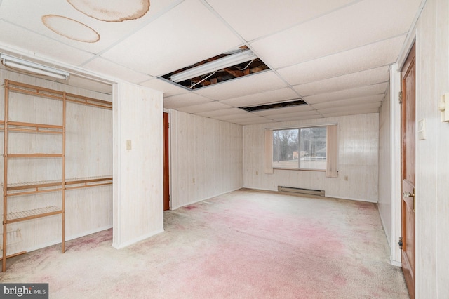 basement with light colored carpet, wooden walls, a drop ceiling, and a baseboard heating unit