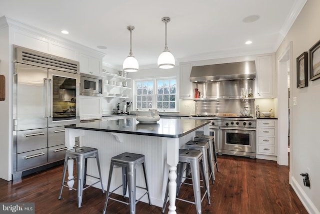 kitchen with a breakfast bar, dark countertops, white cabinets, wall chimney range hood, and built in appliances