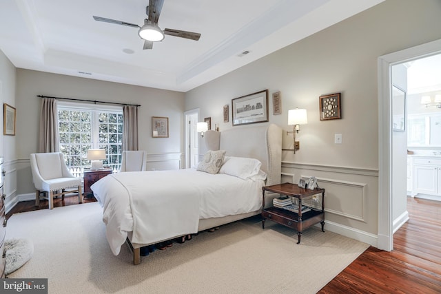 bedroom with a decorative wall, wood finished floors, a ceiling fan, wainscoting, and a tray ceiling